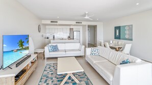 A modern living room with a flat-screen TV, white sofas, a coffee table, and a kitchen in the background.