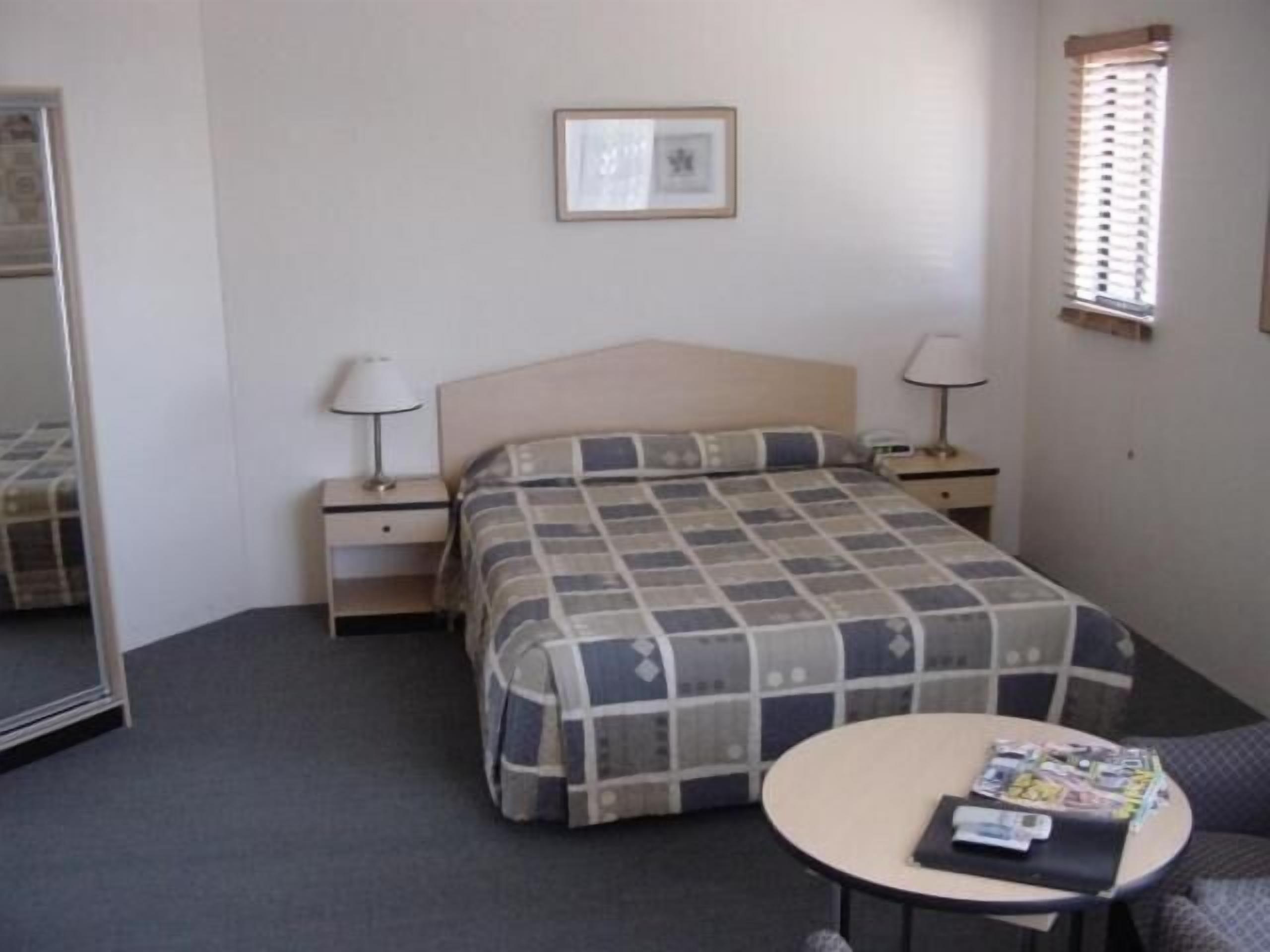 A hotel room with a bed, two bedside tables, a round table with magazines, a chair, and a window with blinds.