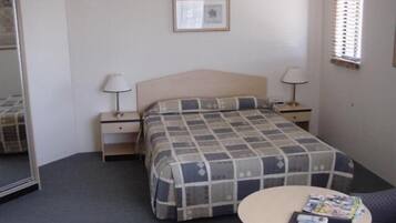 A hotel room with a bed, two bedside tables, a round table with magazines, a chair, and a window with blinds.