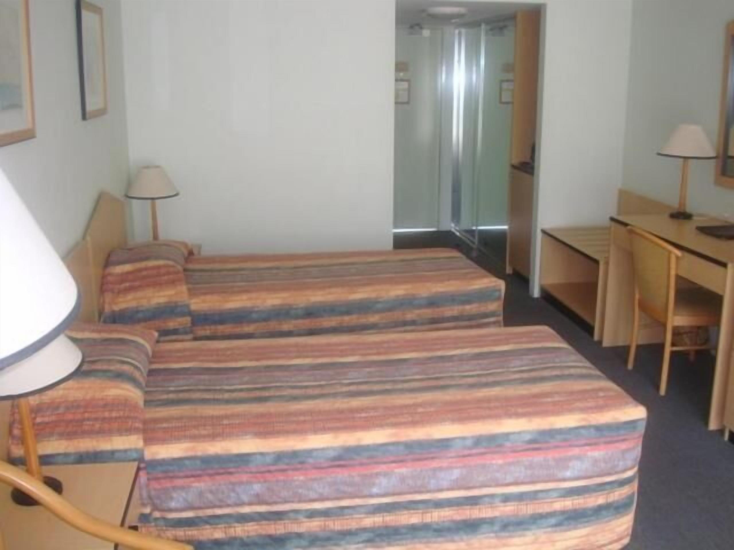 A hotel room with two beds, a desk, a chair, and a lamp.