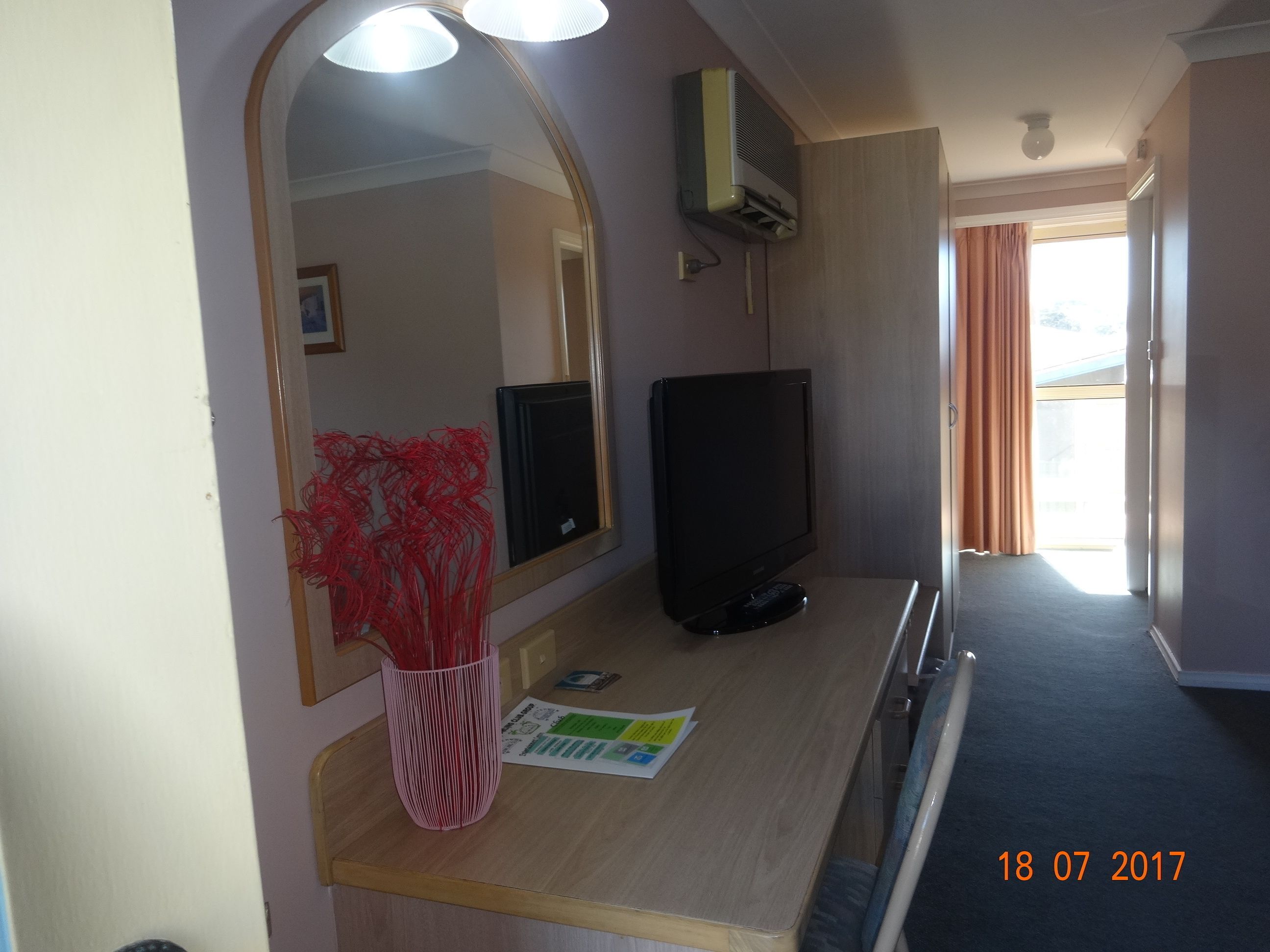 A hotel room with a large mirror, a flat-screen TV, a small table with a vase of red flowers, and a window with curtains.