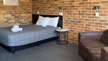 Un dormitorio con pared de ladrillo, una cama con colchón gris y almohadas blancas, una mesita redonda y un sofá marrón.