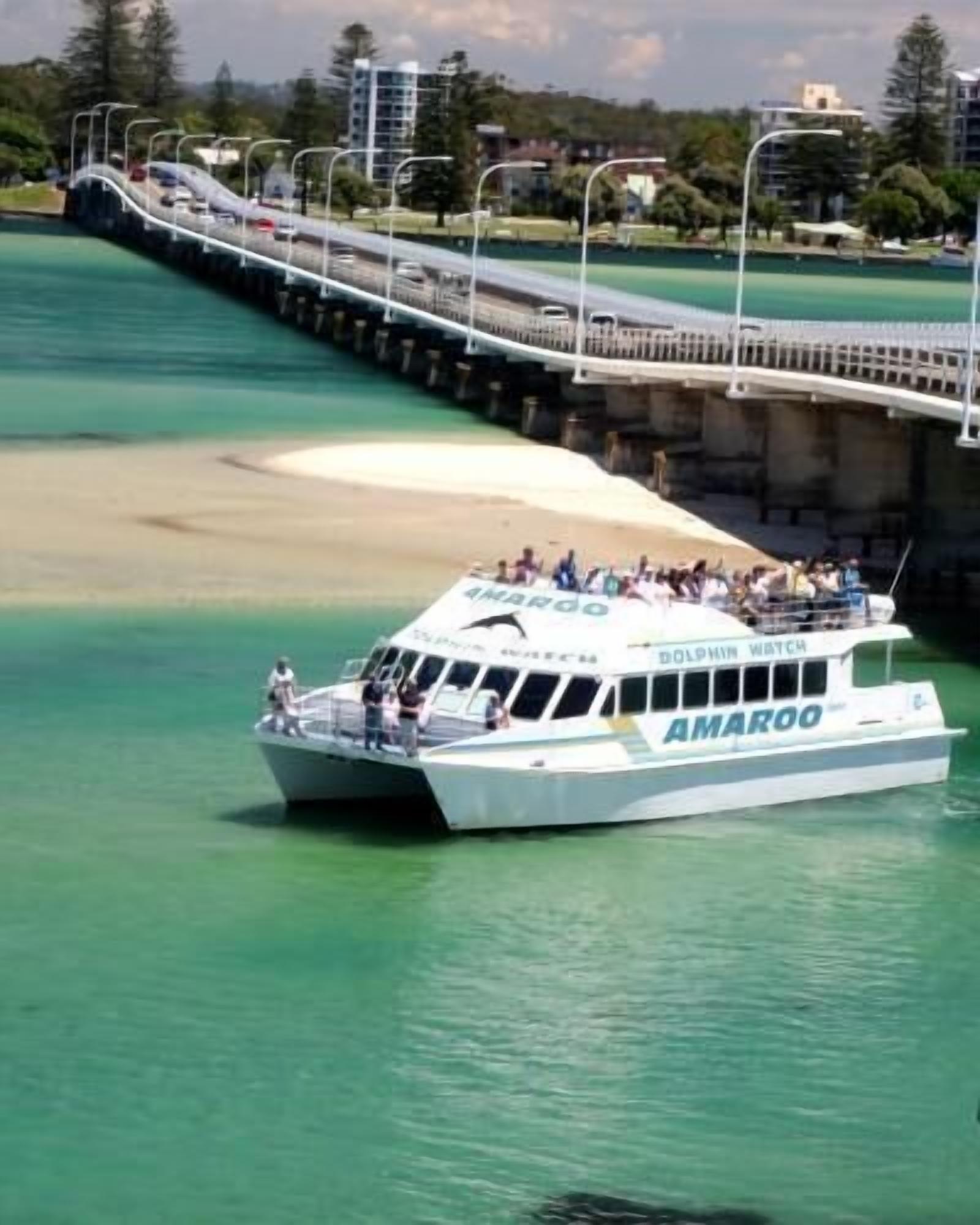 A boat named 'AMAROO' with passengers, docked near a bridge and waterfront.