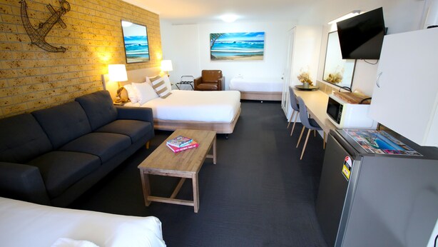 A compact hotel room with a sofa, a bed, a small table with a book, a TV, and a refrigerator. - Tweed Harbour Motor Inn (Tweed Heads)