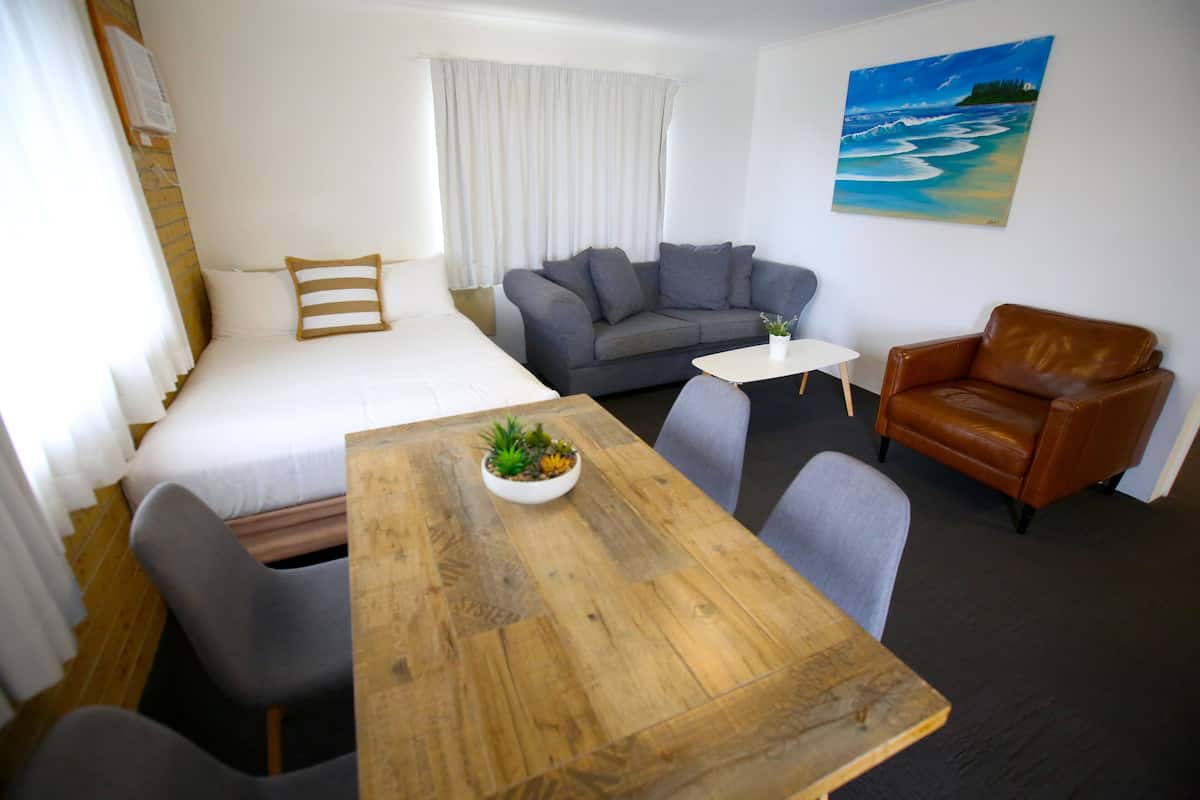 A room with a bed, a grey sofa, a brown leather chair, a wooden table with a plant, and a painting of a beach scene.