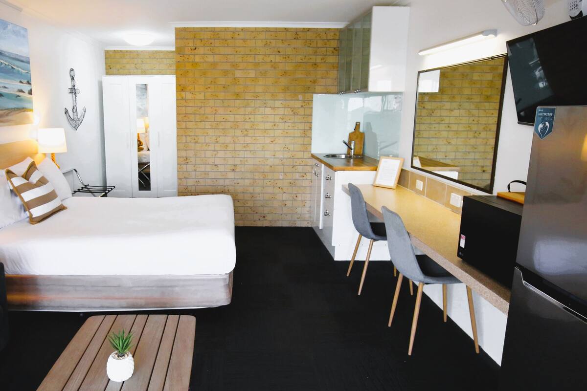 A hotel room with a bed, desk, chairs, a small potted plant, a TV, and a brick wall.