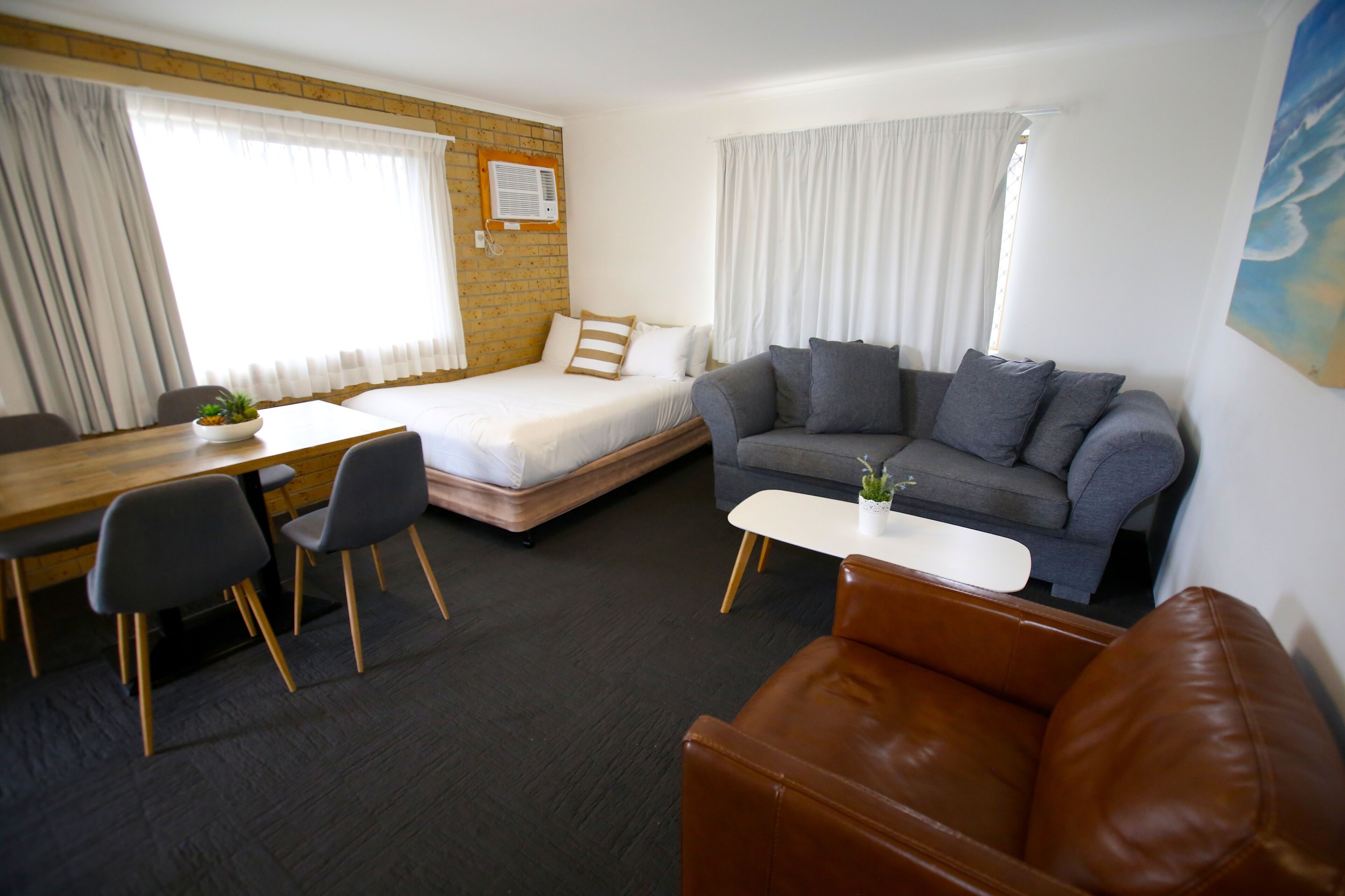 A compact room with a bed, a grey sofa, a brown leather couch, a dining table with chairs, and a small white table with a plant.