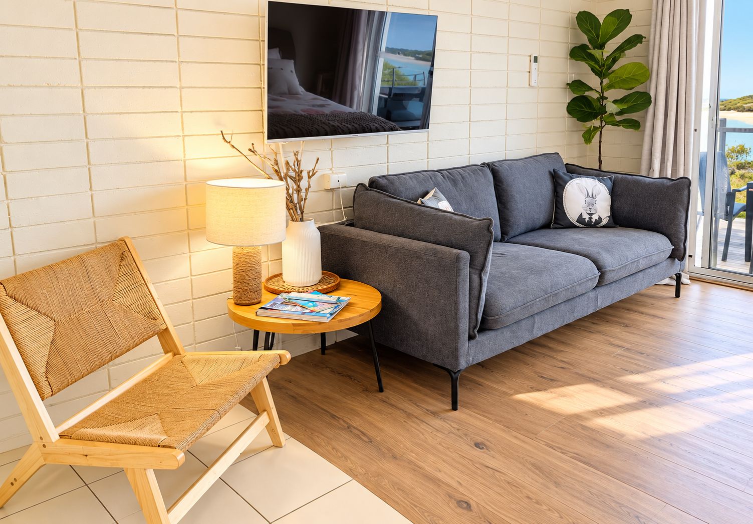 A modern living room with a grey sofa, a wicker chair, a small round table with a lamp, and a TV mounted on the wall.