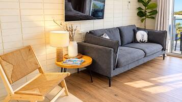 A modern living room with a grey sofa, a wicker chair, a small round table with a lamp, and a TV mounted on the wall.