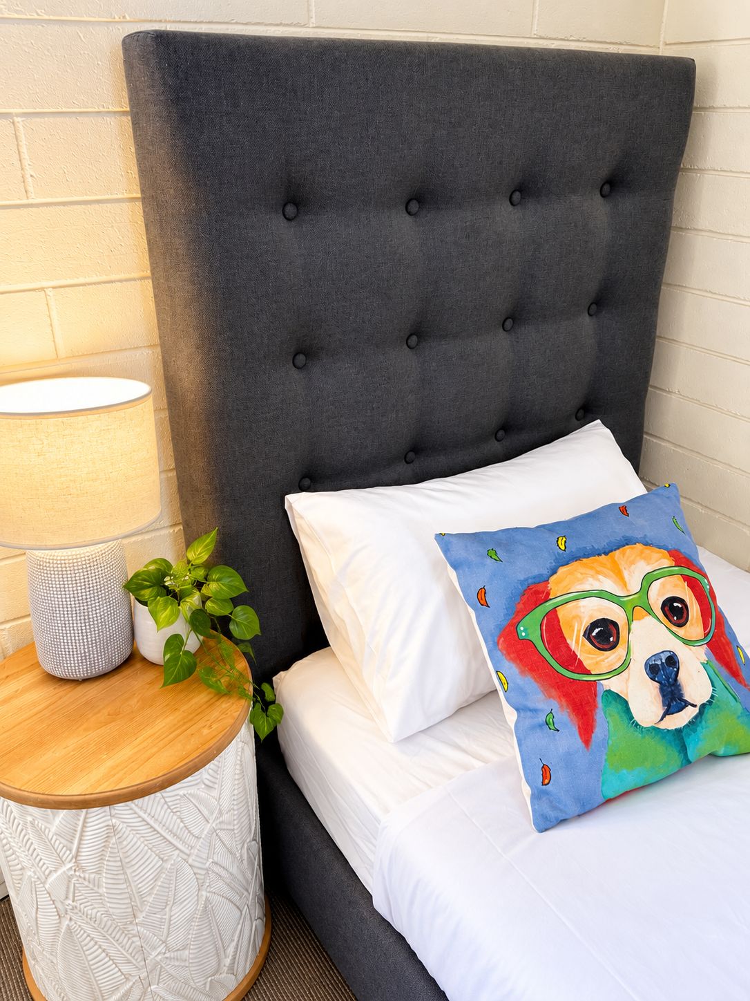 A bed with a tufted headboard, white bedding, and a colorful pillow with a dog illustration.