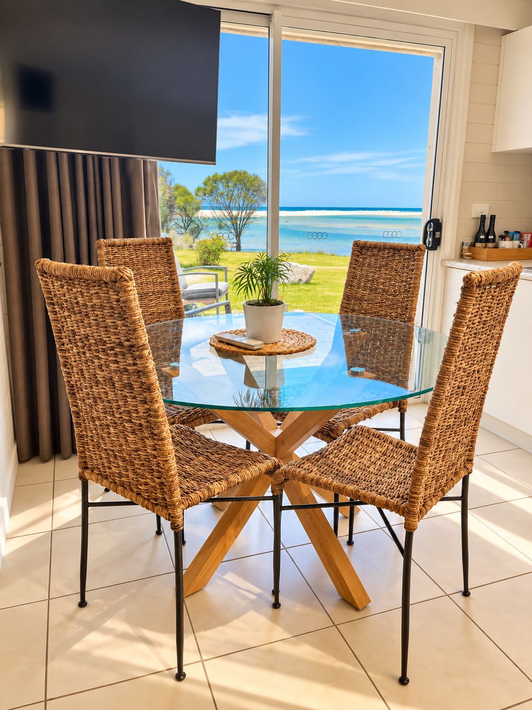 A dining area with a glass table and wicker chairs, overlooking a beach.
