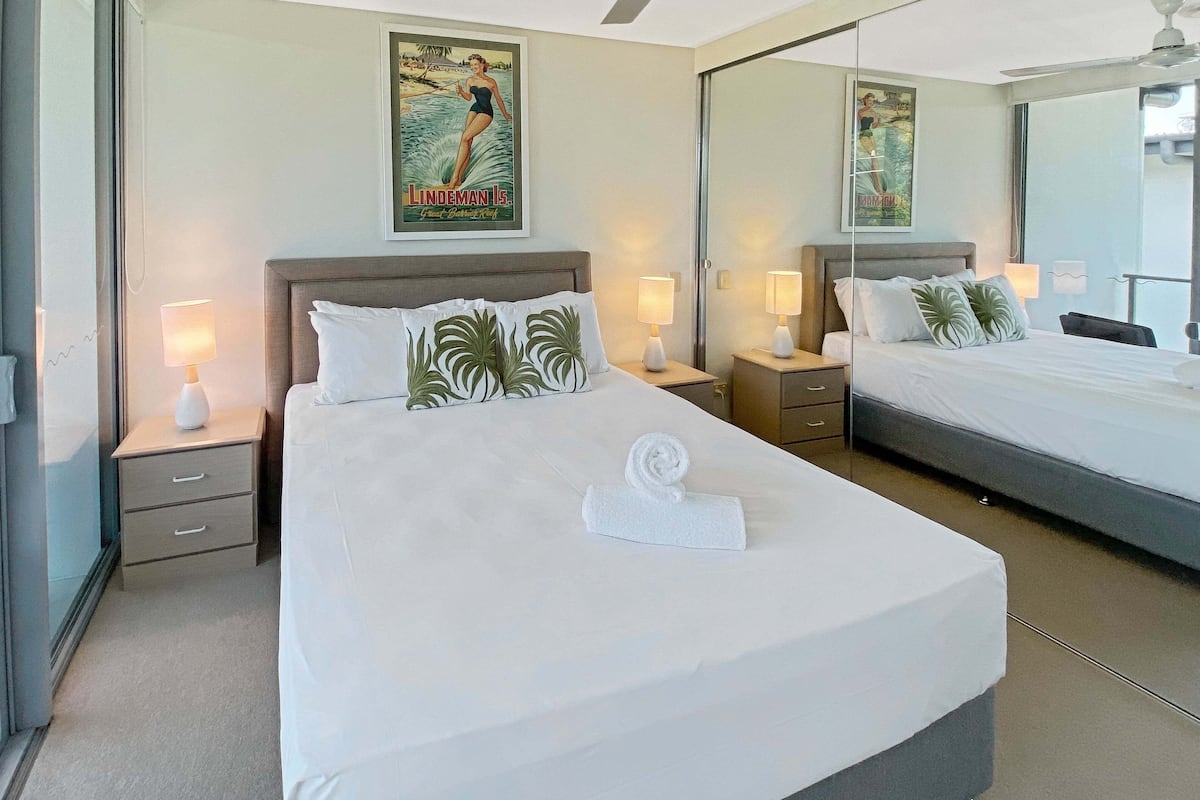 Quarto de hotel com uma cama grande, duas mesas de cabeceira com luminárias, um ventilador de teto, um pôster na parede e um espelho grande refletindo o quarto.