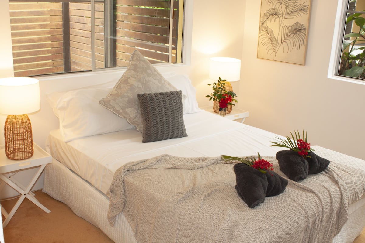 Cama bem arrumada com manta cinza, duas almofadas e duas toalhas com flores. Dois abajures e uma mesinha de cabeceira com um vaso de flores.