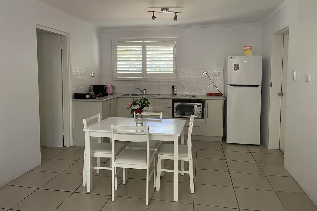 Uma cozinha compacta com armários brancos, uma mesa de jantar com quatro cadeiras, micro-ondas, geladeira e uma janela com persianas.