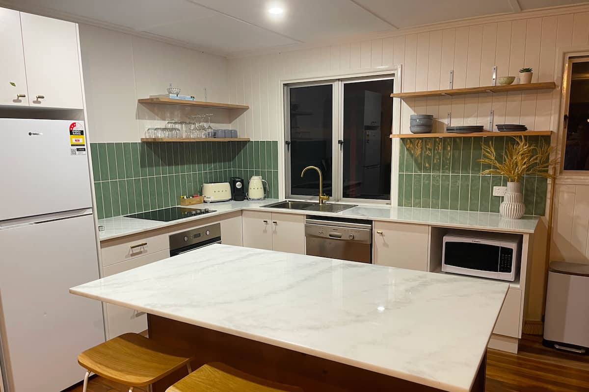 Cozinha moderna com bancada de mármore, banquetas de madeira, lava-louças, micro-ondas e janela com vista.
