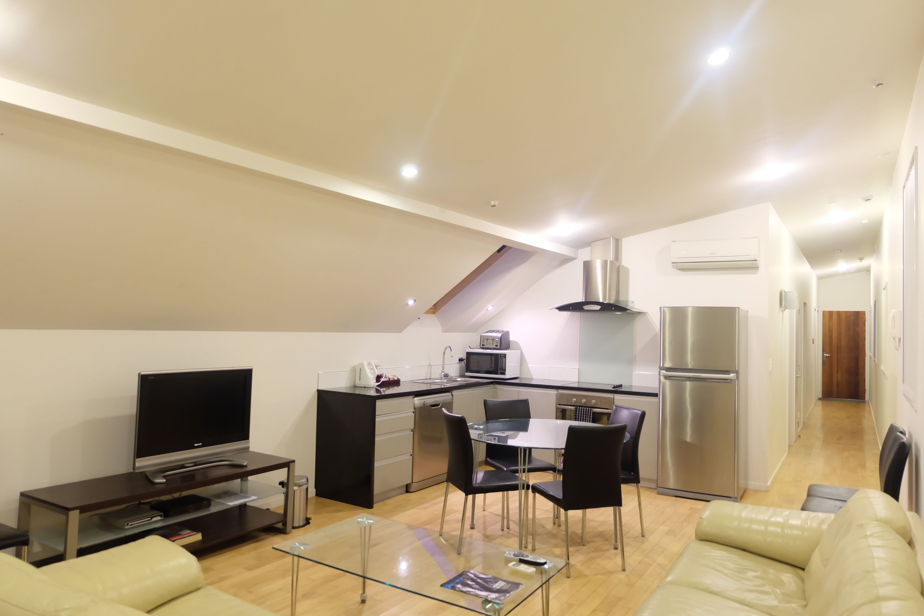 A modern apartment with a living room, a glass coffee table, a TV, a kitchen with a dining area, and a small balcony.