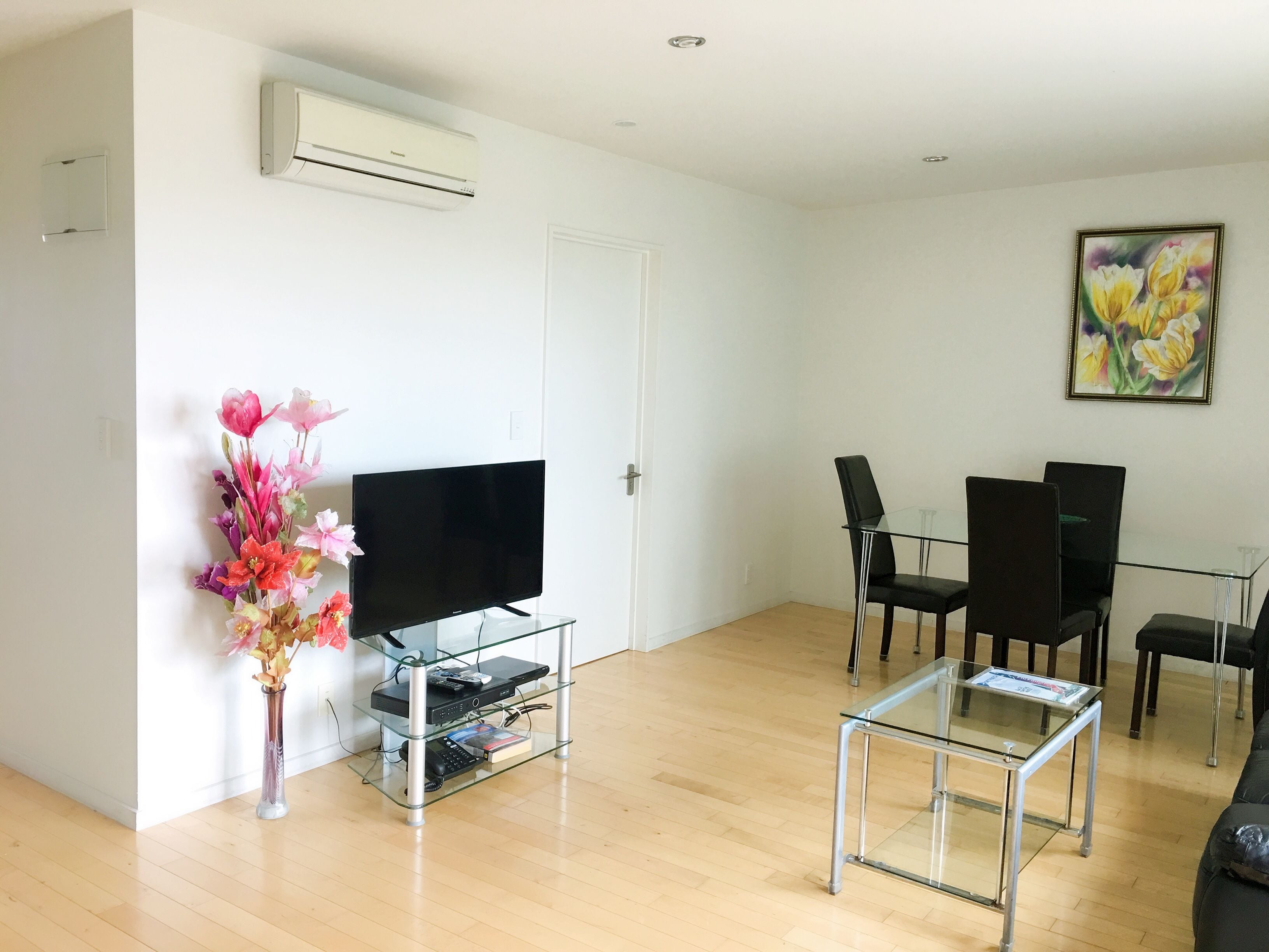 Un salon moderne avec un téléviseur à écran plat posé sur une table en verre, des chaises noires, un plancher en bois et un tableau accroché au mur.