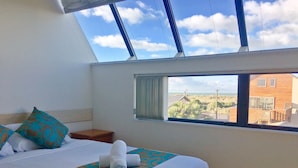 A bedroom with a large bed, a window with a view of the beach, and a skylight. - Beachlife Apartments (Christchurch)