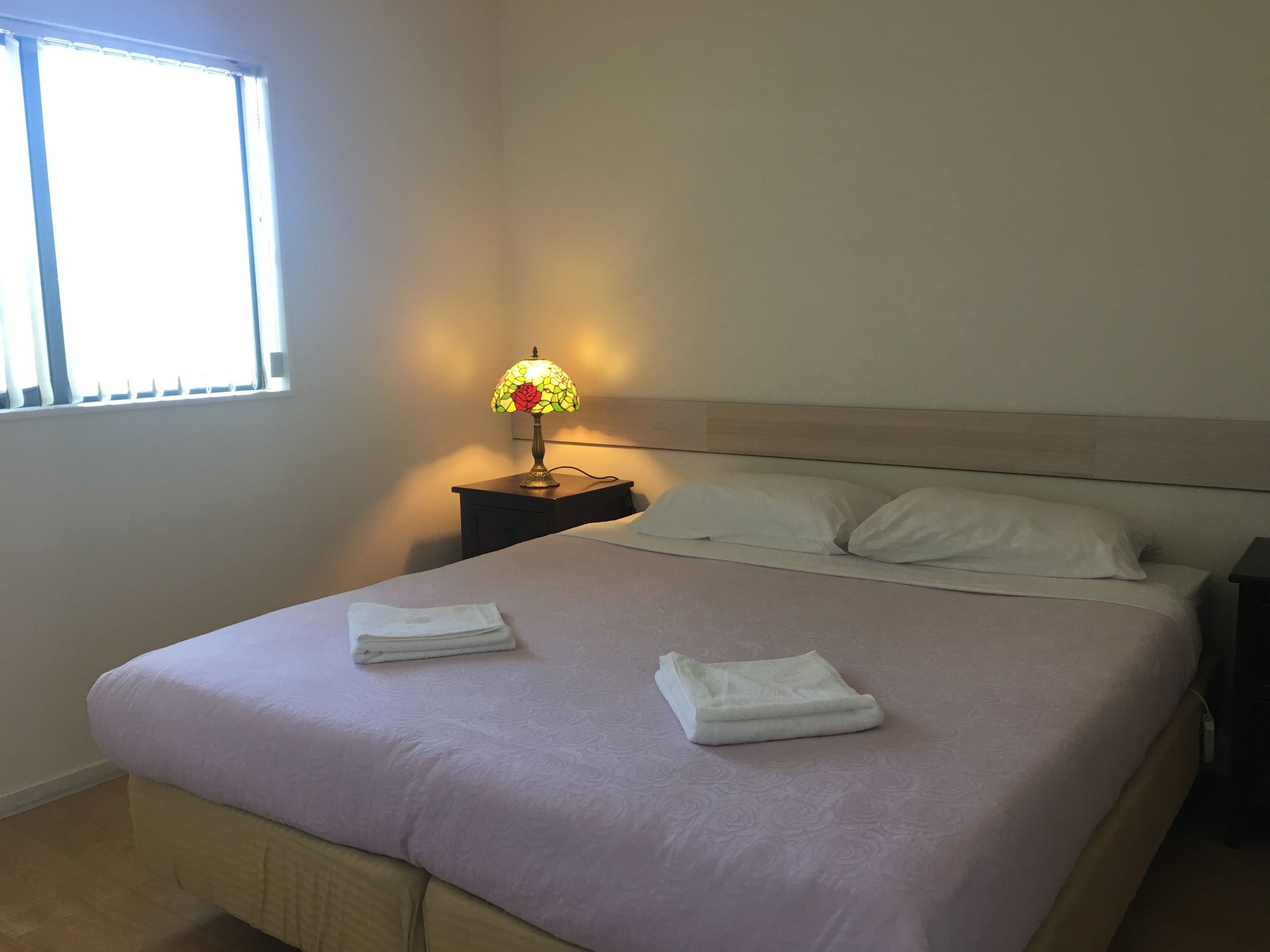 A neatly made bed with a light purple bedspread and two folded white towels. A small table with a lamp and a power outlet is beside the bed.