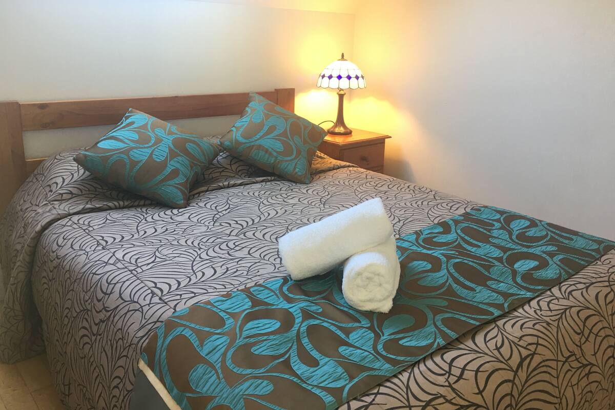 A neatly made bed with patterned bedding, a bedside lamp, and a rolled towel on the bed.