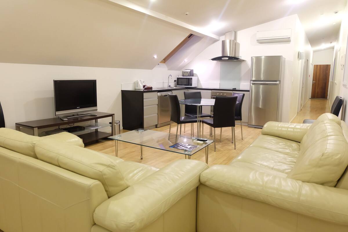 A modern living room with a glass coffee table, beige leather sofa, and a flat-screen TV.