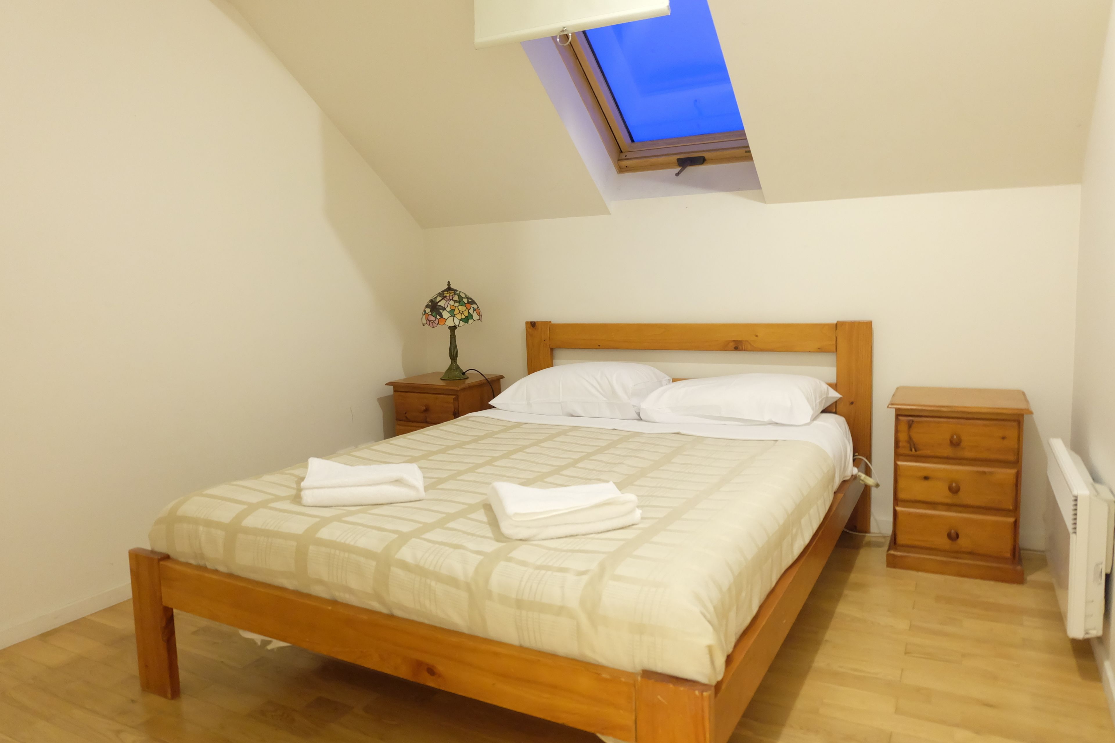 Een slaapkamer met een houten bed, een nachtkastje met een lamp, een kast en een dakraam.