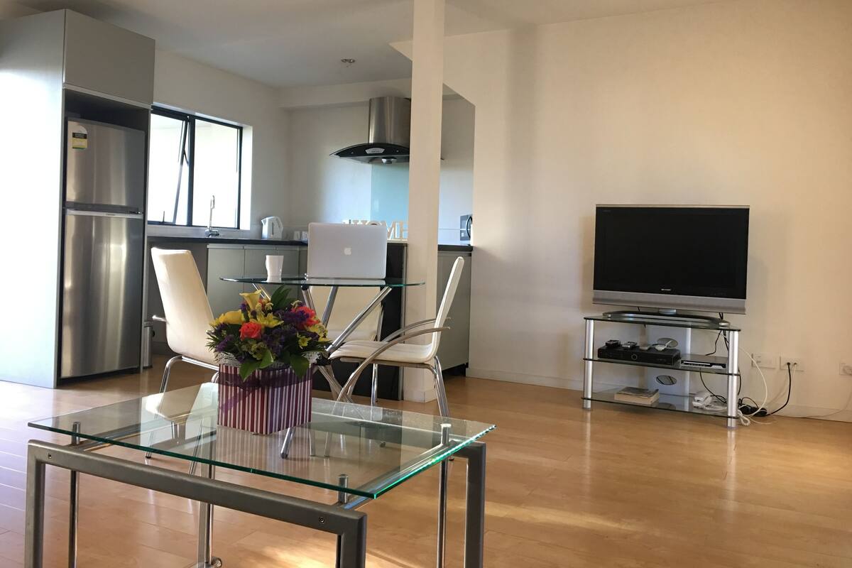 A modern kitchen with stainless steel appliances, a glass dining table, and a flat-screen TV.
