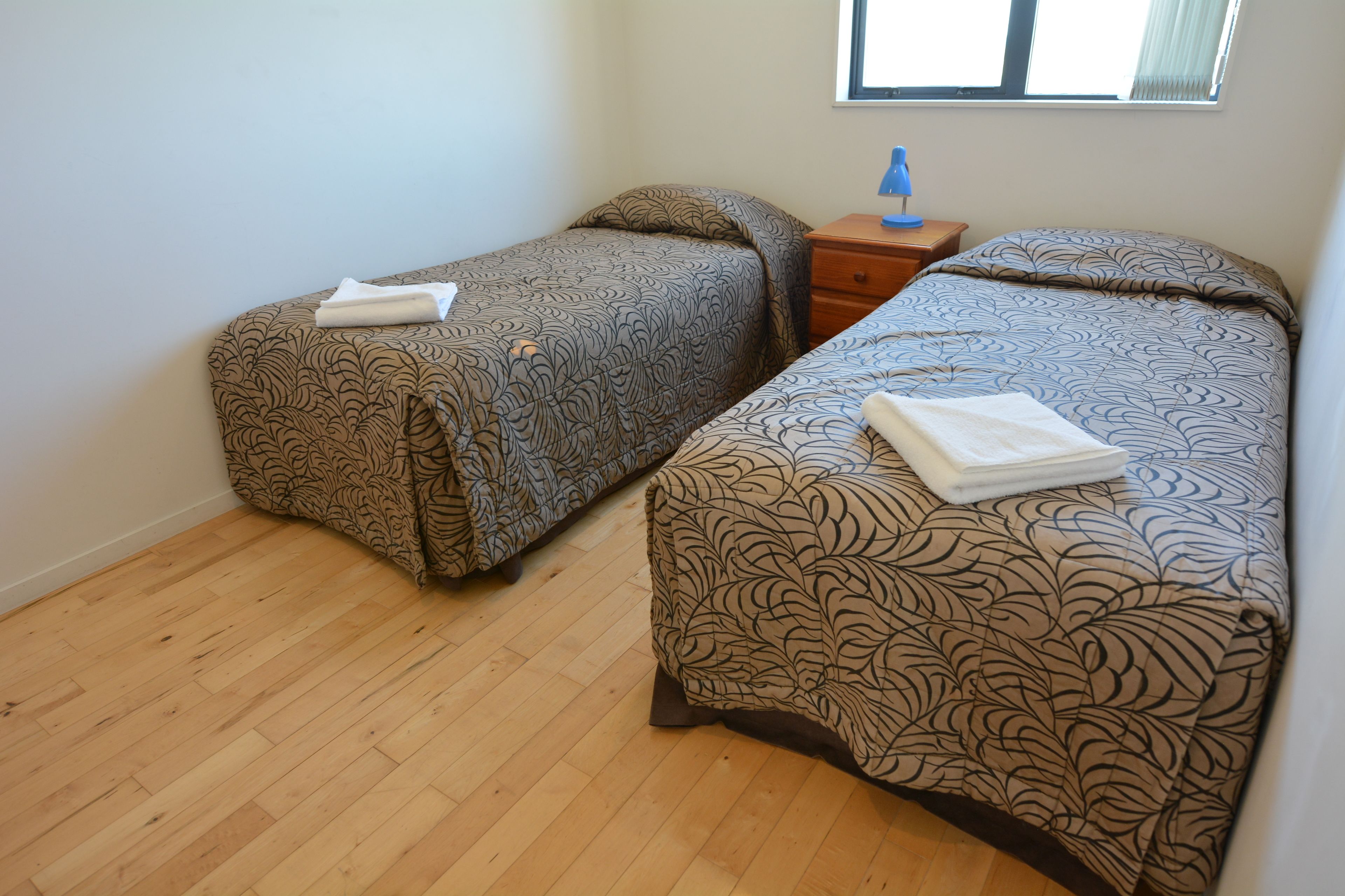 Two single beds with patterned bedding, a wooden nightstand, and a window with blinds.