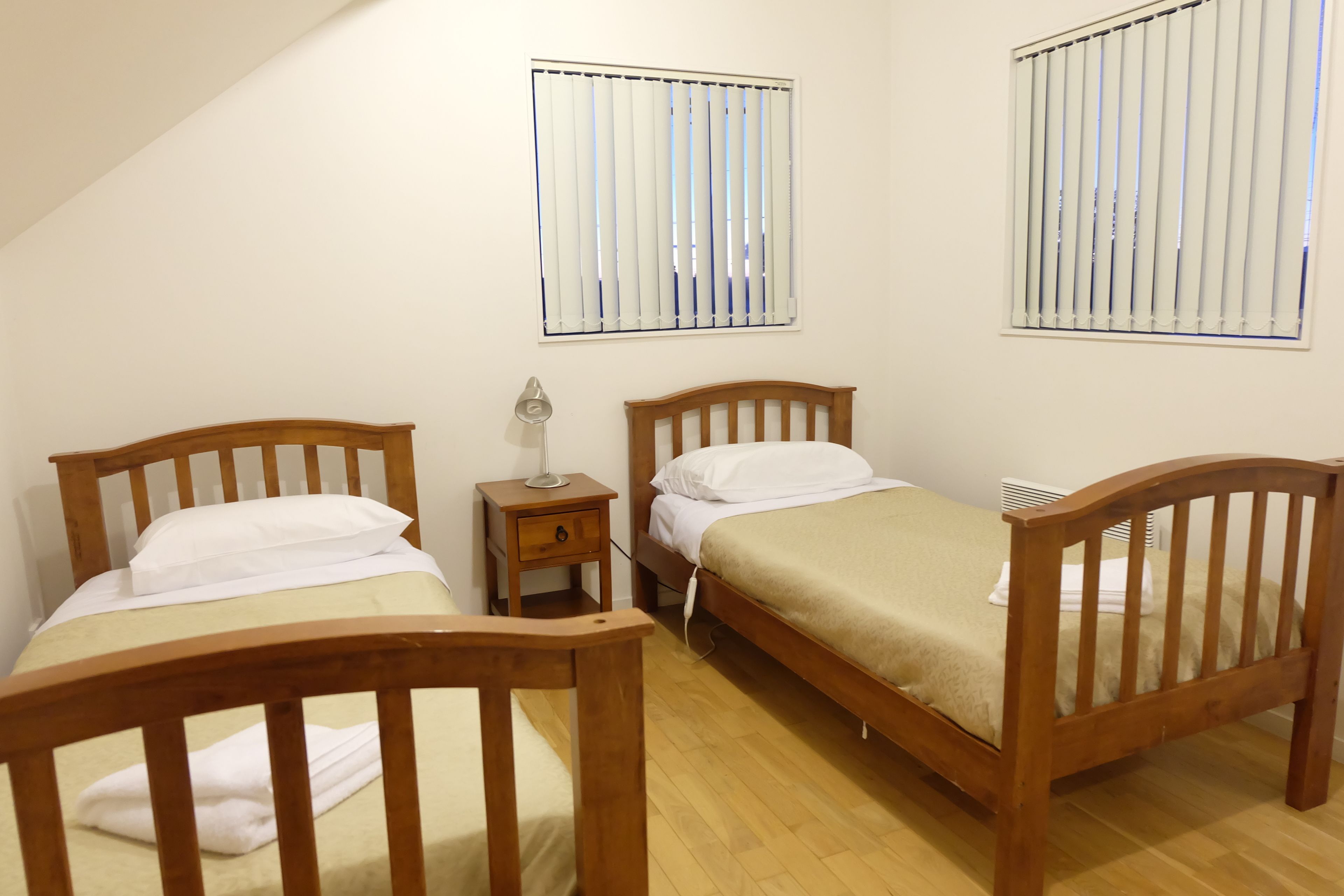 Een kamer met twee houten bedden, een nachtkastje met een lamp en een raam met jaloezieën.