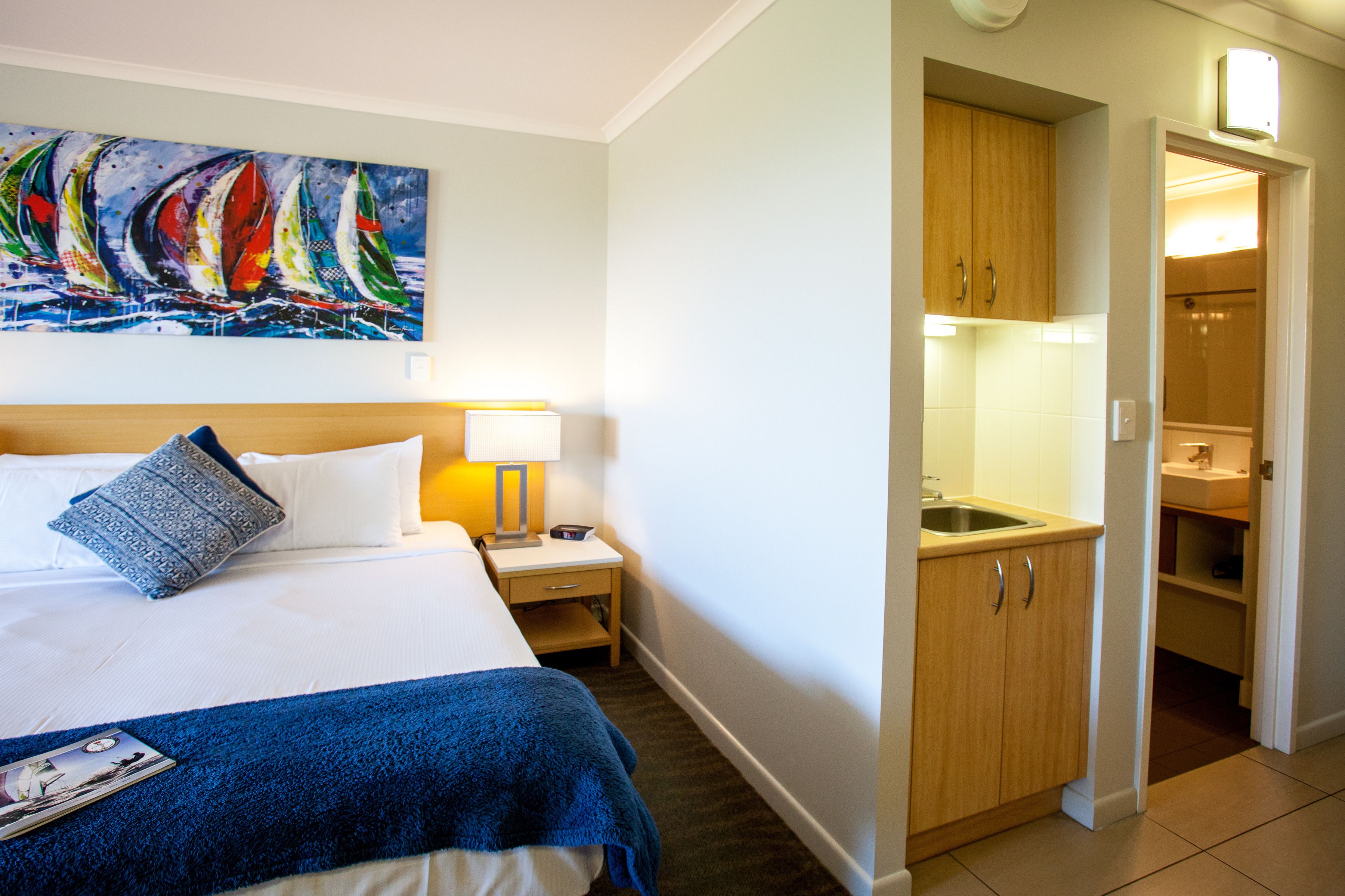 A hotel room with a bed, a nightstand with a lamp, a painting of sailboats, and a view into the bathroom.