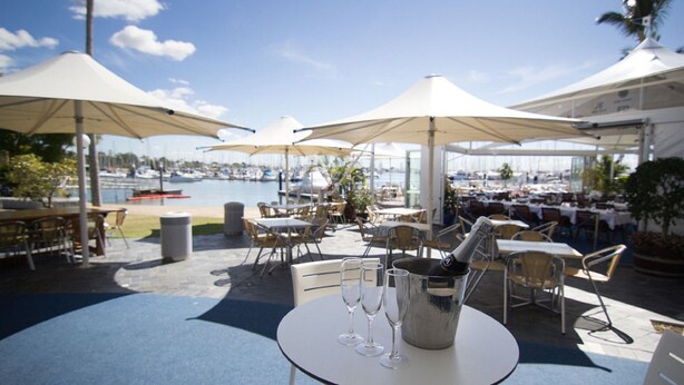 An outdoor dining area with tables and chairs, umbrellas, and a view of the marina. - Manly Marina Cove Motel Brisbane (Manly)