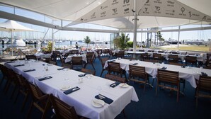 An outdoor dining area with tables set for guests, covered by large umbrellas with logos, and a marina view in the background. - Manly Marina Cove Motel Brisbane (Manly)