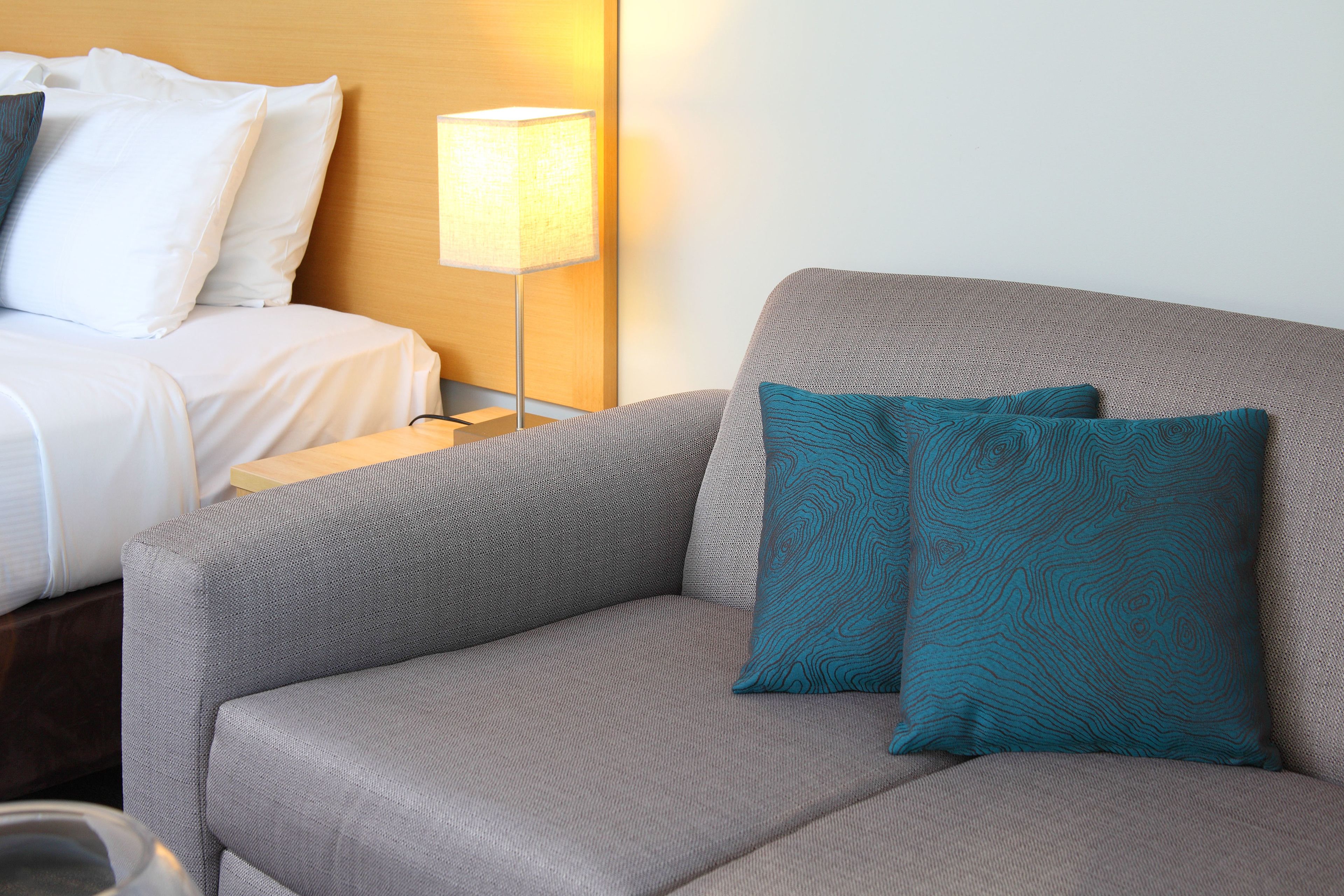Habitación de hotel con un sofá gris, dos almohadas teal estampadas, una mesita de noche con lámpara y una cama con ropa de cama blanca.
