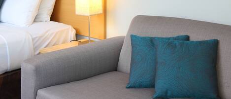 Habitación de hotel con un sofá gris, dos almohadas teal estampadas, una mesita de noche con lámpara y una cama con ropa de cama blanca.