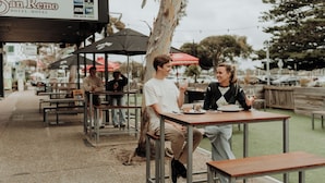 An outdoor dining area with wooden tables and benches, a sign for 'San Remo Hotel-Motel', and a tree in the background. - San Remo Hotel Motel (San Remo)