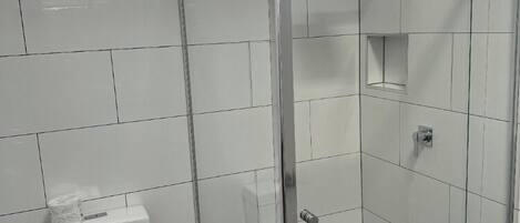 A modern bathroom with white tiles, a toilet, a shower with a glass door, and a window with a view.