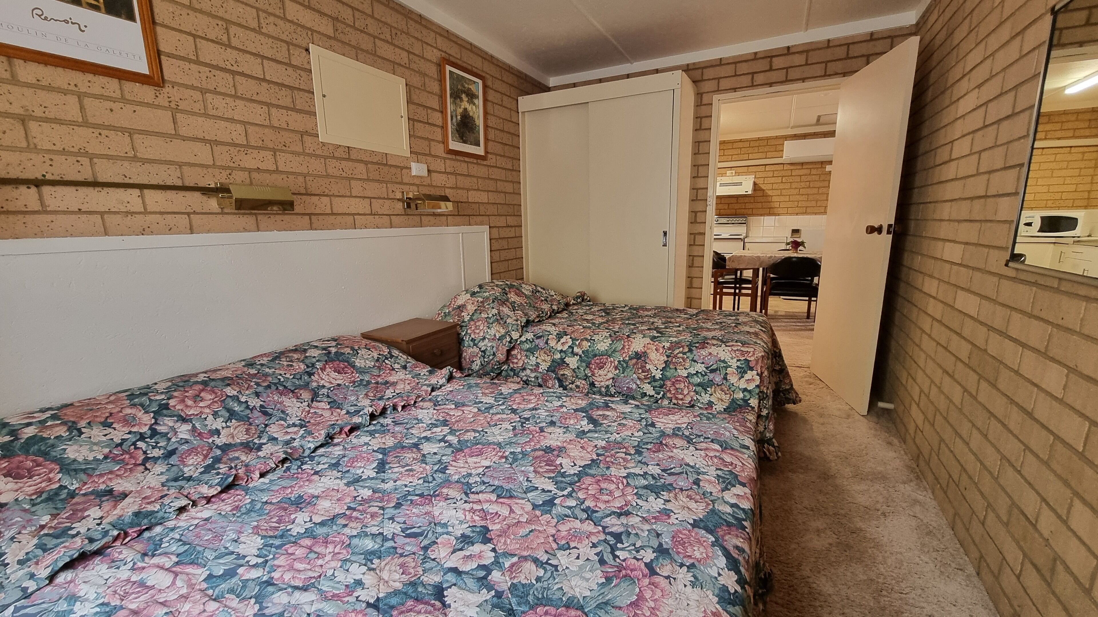 A bedroom with two beds, a brick wall, a door leading to a kitchen, and a framed picture on the wall.