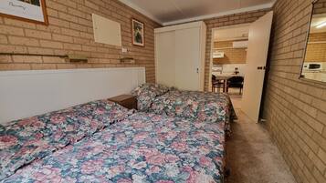 A bedroom with two beds, a brick wall, a door leading to a kitchen, and a framed picture on the wall.
