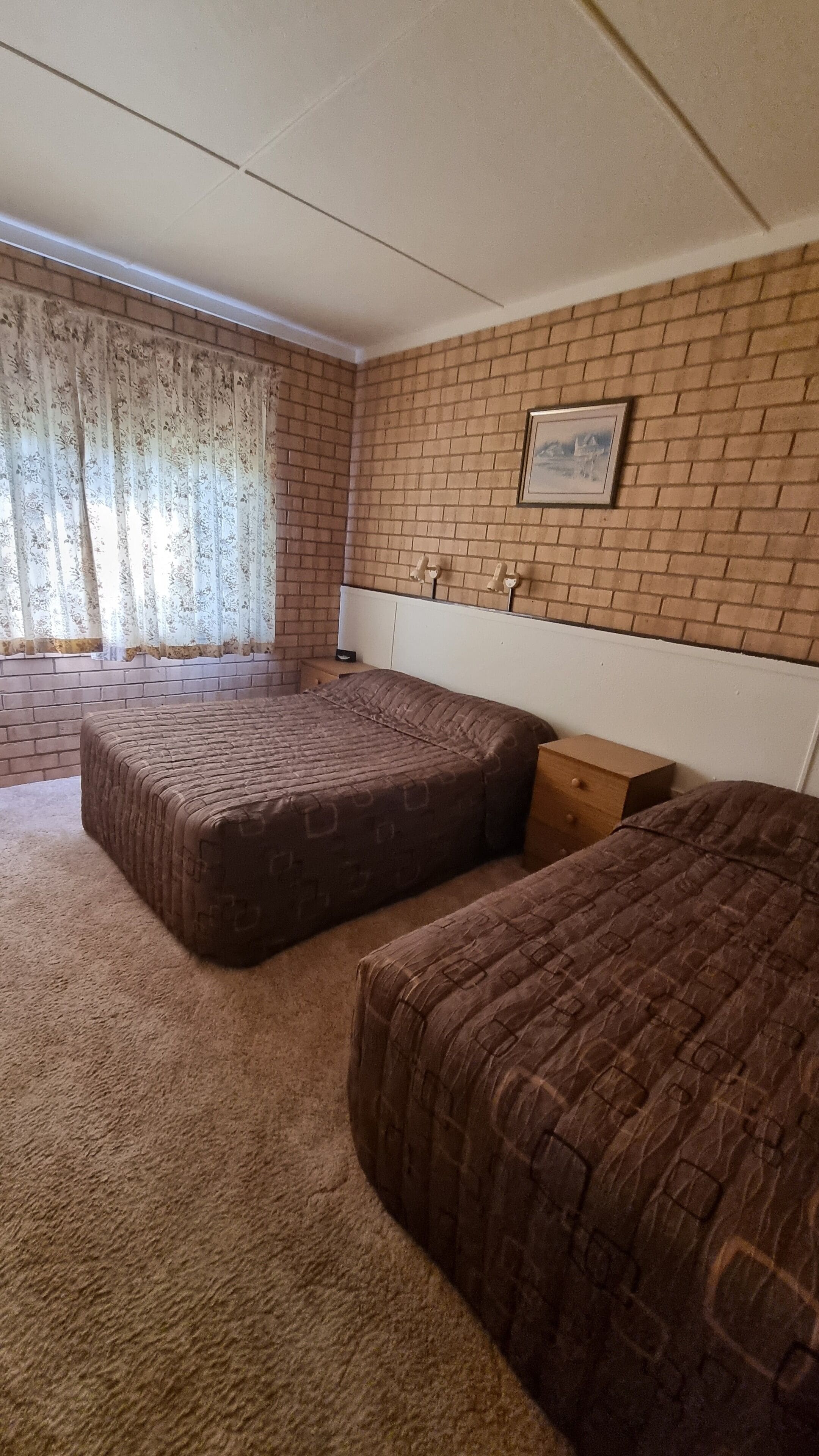 Een kamer met twee bedden, een bakstenen muur, een lijst met een afbeelding en een raam met gordijnen.