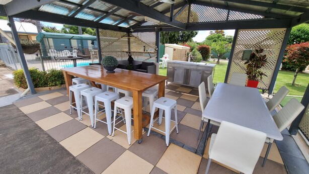Un patio al aire libre con una mesa de madera y taburetes de bar blancos, una parrilla y un área de estar cubierta.