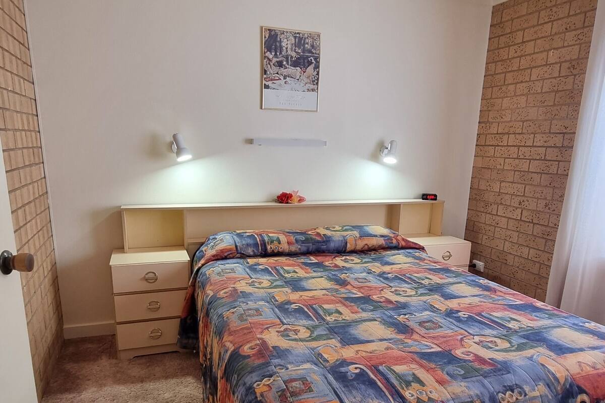 Een slaapkamer met een bed, nachtkastje, dressoir, een wandplank en een ingelijste foto.