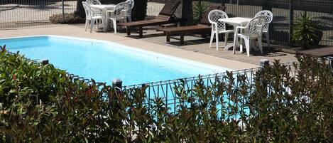 A swimming pool with lounge chairs and tables, surrounded by a fence and greenery.
