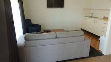 Un saló amb un sofà gris, un butac armchair blau, un televisor muntat a la paret i una cuina amb armaris blancs i un rentapeus.