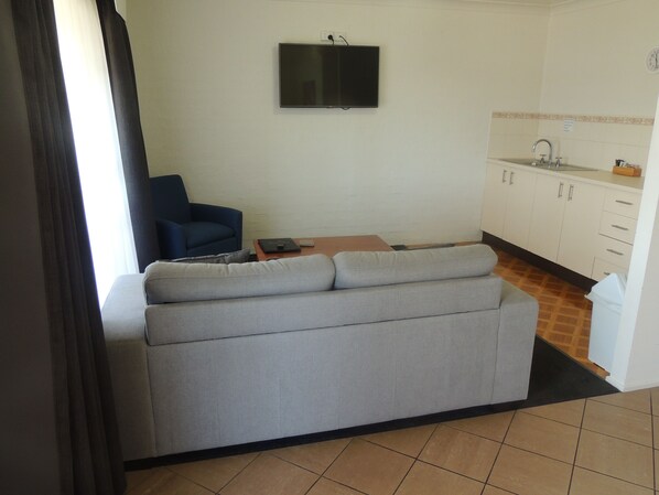 A living room with a grey sofa, a blue armchair, a television mounted on the wall, and a kitchen area with white cabinets and a sink. - Argyle Terrace Motor Inn (Batemans Bay)