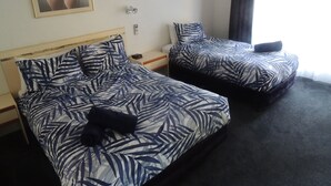 A hotel room with two beds, each with a leaf-patterned bedspread, a nightstand with a clock, a painting on the wall, and a window with curtains.