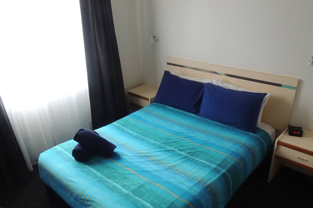 Una habitación de hotel con cama, mesita de noche, lámpara y ventana con cortinas.
