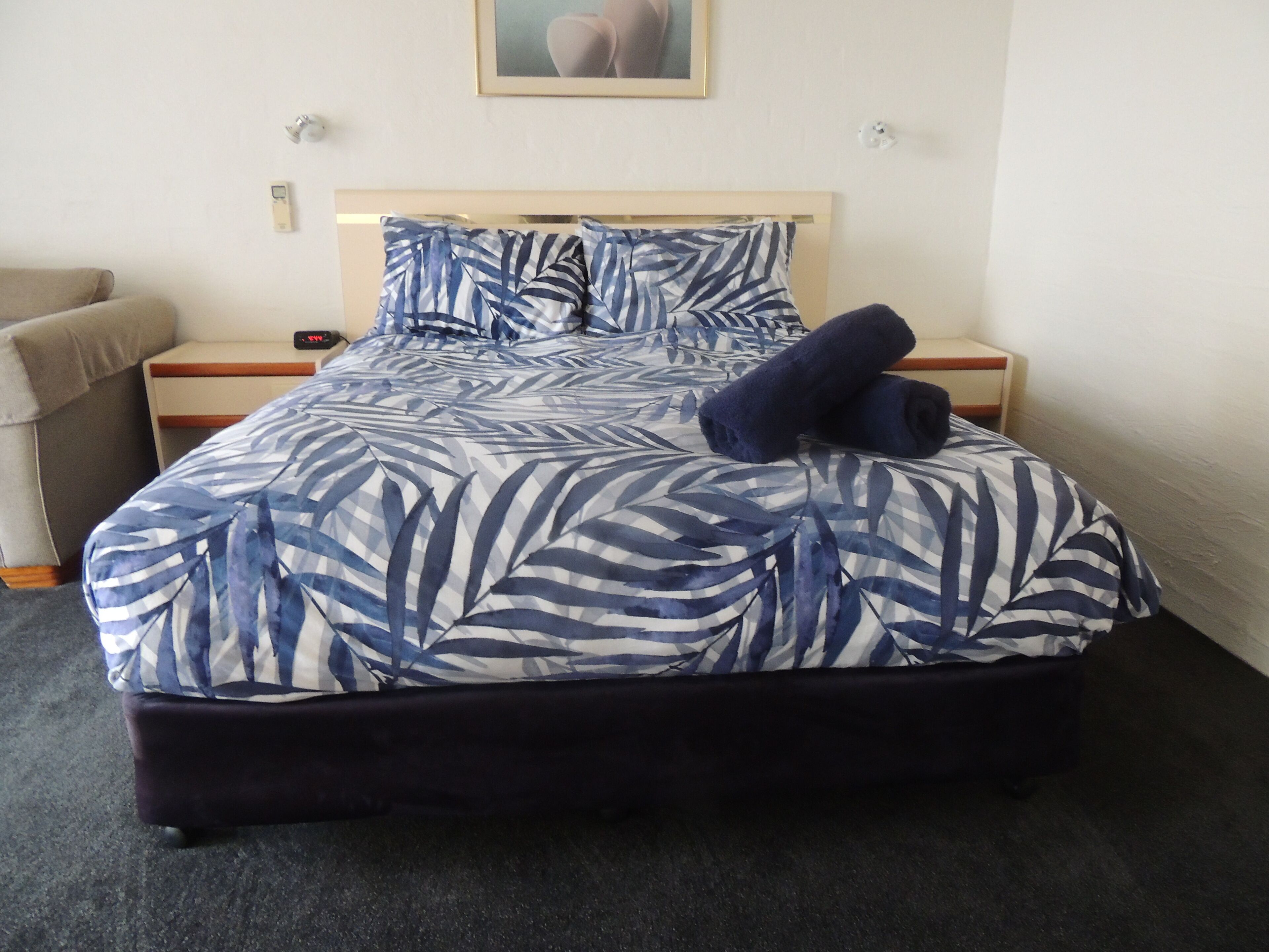 A bed with a blue and white leaf pattern bedspread, two dark blue pillows, a wooden headboard, and a small bedside table with a lamp.