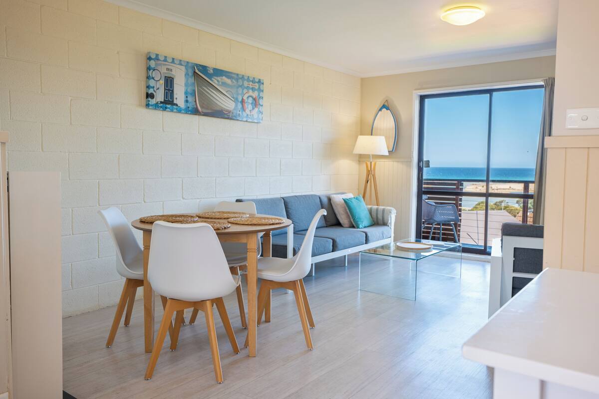 A room with a dining table, chairs, a sofa, a lamp, and a view of the beach through a large window.