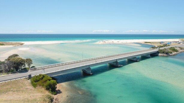 Un puente sobre una masa de agua azul cristalina, con una playa y vegetación a los lados.