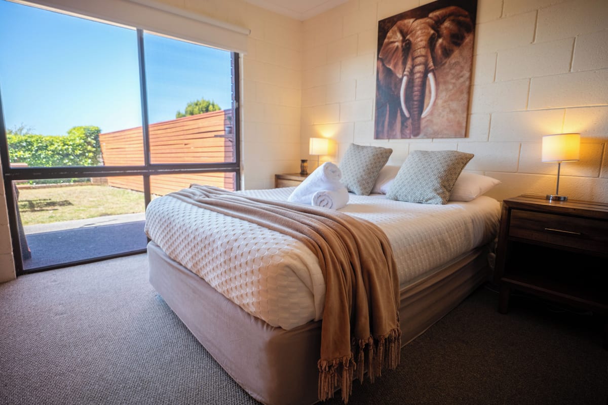 A bedroom with a large bed, two bedside tables with lamps, a large window, and a wall-mounted elephant artwork.